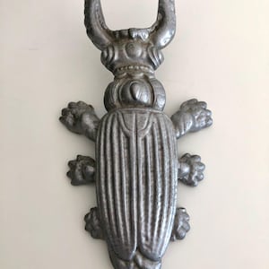 May include: A silver-colored metal decorative beetle with large curved horns and detailed body segments. The beetle has six legs and is likely a decorative item or wall hanging.
