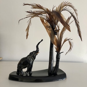 May include: A black carved elephant figurine standing under a black carved palm tree with brown feathers. The figurine is on a black base.