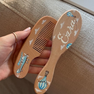 May include: Wooden baby hairbrush and comb set. The brush is painted with the name "Ezra" in cursive, along with blue hot air balloons and clouds. The comb features a blue airplane design and white clouds. The set is made of light-colored wood.