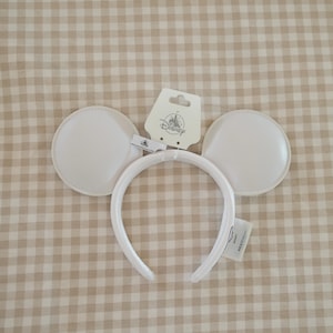 Puede incluir: Diadema blanca con orejas de Mickey Mouse y etiqueta de Disney. La diadema presenta dos orejas grandes y redondas y una banda curva. Las orejas son de material liso. La etiqueta de Disney es blanca con texto negro y un logotipo de castillo.