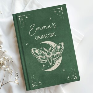 May include: A dark green book with the title "Emma's Grimoire" in elegant script. The cover features a moth design set against a crescent moon, with stars and decorative corner accents. The book is set on a white surface.