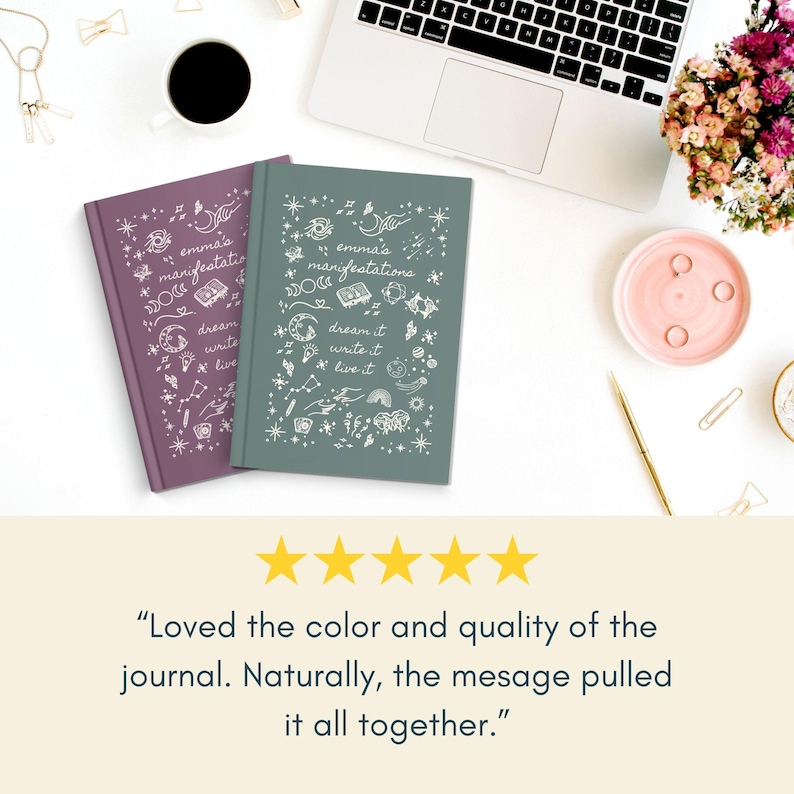 May include: Two hardback journals, one in dusty rose and one teal, are shown on a white surface. Each journal has the text "emma's manifestations" and the words "dream it, write it, live it" with decorative doodles. A laptop, coffee cup, and flowers are also visible.