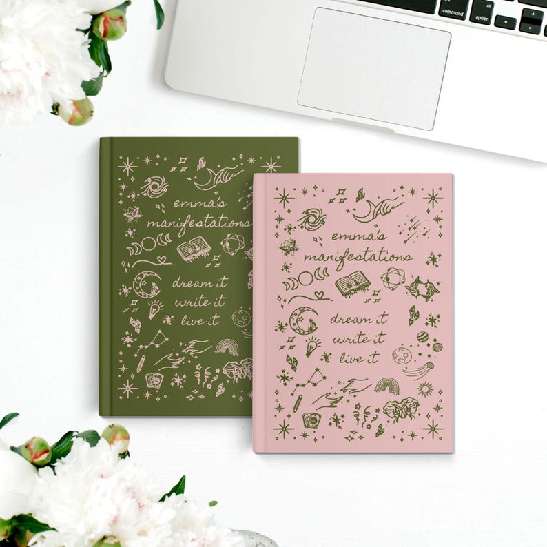 May include: Two hardback journals, one in olive green and the other in pink, are shown on a white surface. The journals have the text "emma's manifestations" and the phrases "dream it, write it, live it" with celestial illustrations.