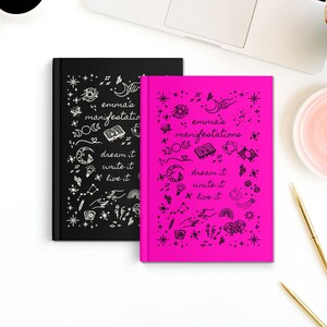 May include: Two hardcover journals, one black and one hot pink, are shown on a white surface. Both journals feature the text "emma's manifestations" and the phrases "dream it, write it, live it" with celestial and whimsical illustrations.