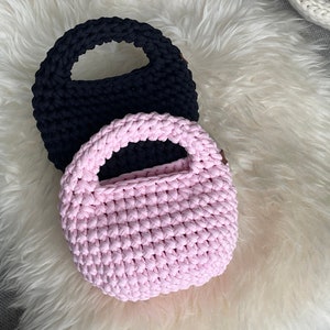 Könnte beinhalten: Zwei runde, gehäkelte Handtaschen mit integrierten kreisförmigen Griffen. Eine Tasche ist einfarbig schwarz, die andere einfarbig hellrosa. Beide Artikel zeichnen sich durch eine dicke, strukturierte Garnkonstruktion mit einem gleichmäßigen Maschenbild aus.