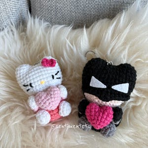 May include: Two handmade crochet keychains. One is a white cat with a pink bow and heart. The other is a black superhero with a pink heart. Both keychains are on a fluffy white surface.