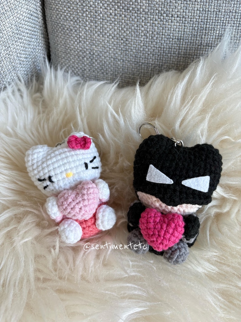 May include: Two handmade crochet keychains. One is a white cat with a pink bow and heart. The other is a black superhero with a pink heart. Both keychains are on a fluffy white surface.