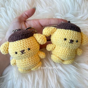 以下が含まれることがあります： 茶色のベレー帽をかぶった、手作りの黄色の犬の編みぐるみキーホルダーが2つ。各キーホルダーには黒い目と小さな黒い鼻があります。キーホルダーはふわふわの白い表面に表示されています。