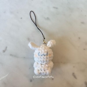 May include: A white crochet bunny charm with blue stitched eyes and blush-colored cheeks. The charm has a black string and silver-colored clasp. The text "@sentimentetc" is visible on the bottom of the charm.