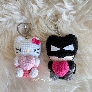 May include: Two handmade crochet keychains. One is white with a pink bow and heart, resembling a cartoon cat. The other is black with white accents and a pink heart. Both have silver key rings. The text "@sentimentetc" is visible.