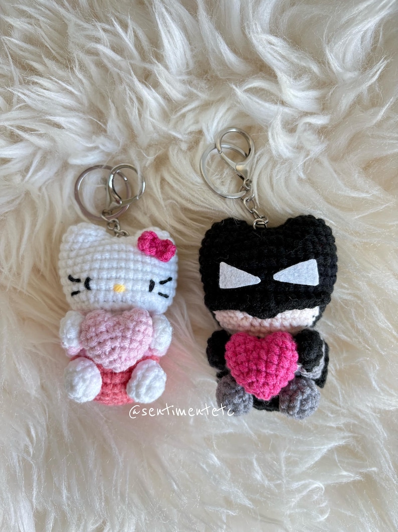 May include: Two handmade crochet keychains. One is white with a pink bow and heart, resembling a cartoon cat. The other is black with white accents and a pink heart. Both have silver key rings. The text "@sentimentetc" is visible.