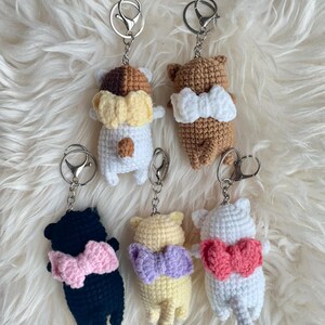 May include: Five crocheted cat keychains with bows. The cats are in different colors: white, brown, black, yellow, and light pink. Each cat has a silver keychain attached.