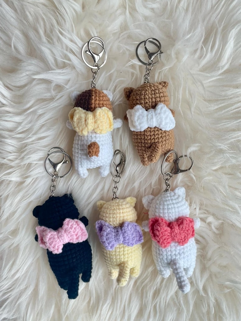 May include: Five crocheted cat keychains with bows. The cats are in different colors: white, brown, black, yellow, and light pink. Each cat has a silver keychain attached.