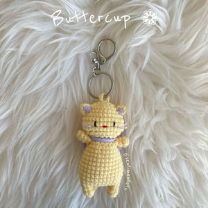 May include: A crocheted yellow cat keychain with purple ears and a red nose. The keychain is attached to a silver key ring.