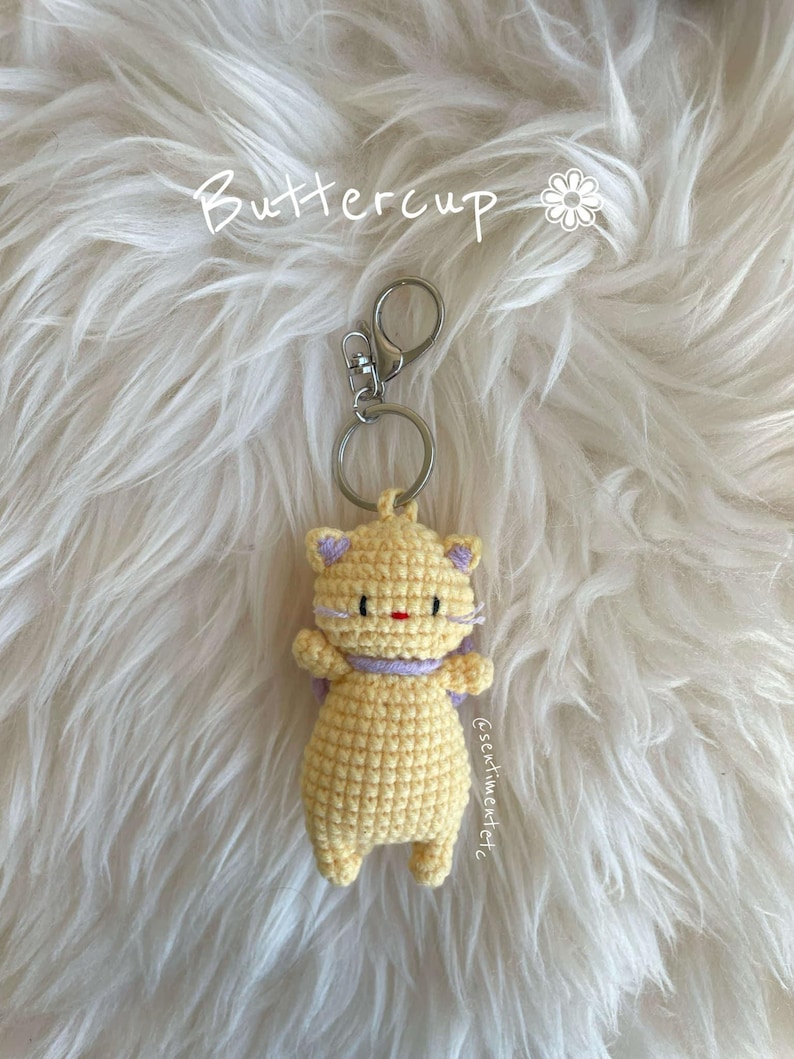 May include: A crocheted yellow cat keychain with purple ears and a red nose. The keychain is attached to a silver key ring.