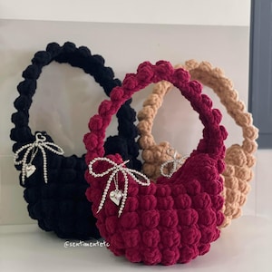 May include: Three crocheted handbags in black, burgundy and beige. Each bag has a heart-shaped charm attached to the handle.