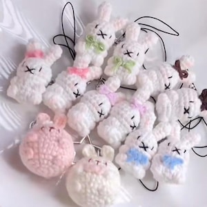 May include: A collection of handmade crochet bunny keychains in various colors. The bunnies are primarily white with accents of pink, blue, green, and brown. Each bunny has a stitched face and a small bow. The keychains are arranged on a white plate.