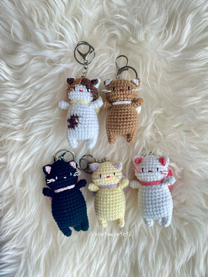 May include: Five crocheted cat keychains in different colors, including black, white, brown, yellow, and calico. Each keychain has a small, white face with black eyes and a pink nose. The keychains are hanging from silver key rings.