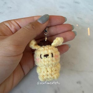 May include: A handmade, yellow crochet keychain in the shape of a cartoon dog with a brown hat. The dog has pink cheeks and a black nose and eyes. The keychain is attached to a silver-colored metal ring. The text "@sentimentetc" is visible.