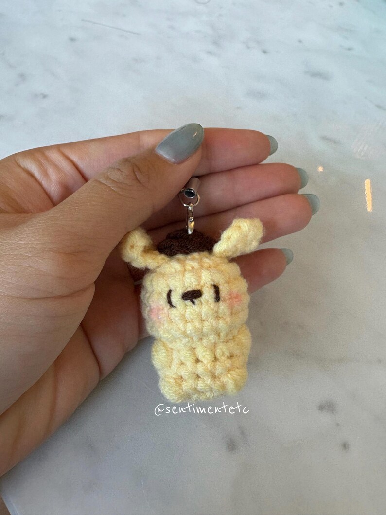 May include: A handmade, yellow crochet keychain in the shape of a cartoon dog with a brown hat. The dog has pink cheeks and a black nose and eyes. The keychain is attached to a silver-colored metal ring. The text "@sentimentetc" is visible.