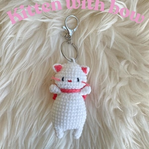 Puede incluir: Llavero de gatito blanco tejido a crochet con un lazo rosa. El llavero está unido a un anillo de llave plateado. El gatito lleva una bufanda rosa. El texto "PATTERN" está escrito debajo del gatito.
