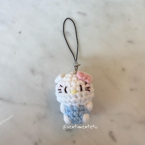May include: A handmade crochet charm featuring a white character with a pink bow, black eyes, and a yellow nose. The character wears a blue outfit. The charm has a black string and a silver clasp. The text "@sentimentetc" is visible.