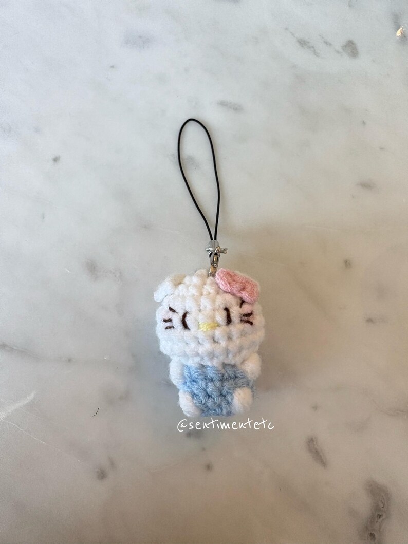 May include: A handmade crochet charm featuring a white character with a pink bow, black eyes, and a yellow nose. The character wears a blue outfit. The charm has a black string and a silver clasp. The text "@sentimentetc" is visible.