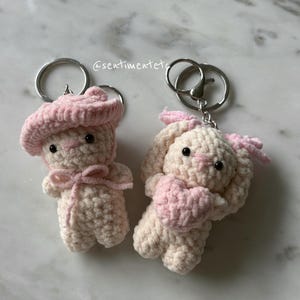 May include: Two handmade crochet keychains. One is a bear wearing a pink beret and bow. The other is a bunny holding a pink heart. Both are cream-colored with pink accents and attached to silver keyrings.