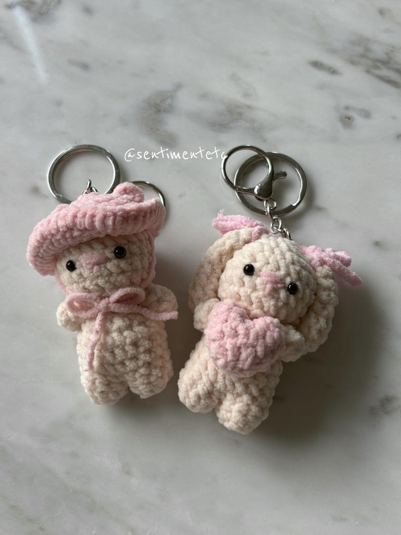 May include: Two handmade crochet keychains. One is a bear wearing a pink beret and bow. The other is a bunny holding a pink heart. Both are cream-colored with pink accents and attached to silver keyrings.
