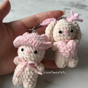 May include: Two handmade crochet bunny keychains. One bunny wears a pink hat and bow, the other holds a pink heart. Both are off-white with black eyes and pink accents. The keychains are attached to silver metal rings.