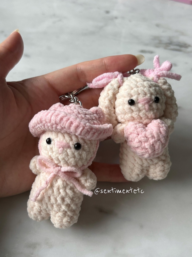 May include: Two handmade crochet bunny keychains. One bunny wears a pink hat and bow, the other holds a pink heart. Both are off-white with black eyes and pink accents. The keychains are attached to silver metal rings.