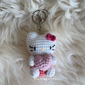 May include: A handmade crochet keychain featuring a white cat-like character with a pink bow and heart. The keychain has black eyes and whiskers, and a silver metal clasp. The text "@sentimentete" is visible on the bottom right.