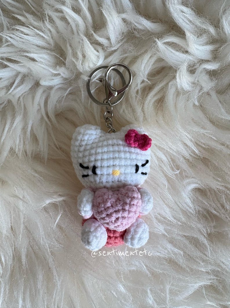 May include: A handmade crochet keychain featuring a white cat-like character with a pink bow and heart. The keychain has black eyes and whiskers, and a silver metal clasp. The text "@sentimentete" is visible on the bottom right.