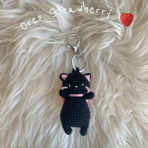 May include: Black crocheted cat keychain with a pink bow, hanging from a silver key ring. The keychain is labeled "Oreo Strawberry" and is on a white fluffy surface.