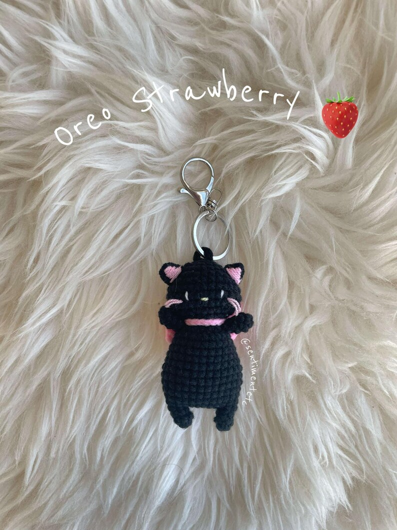 May include: Black crocheted cat keychain with a pink bow, hanging from a silver key ring. The keychain is labeled "Oreo Strawberry" and is on a white fluffy surface.