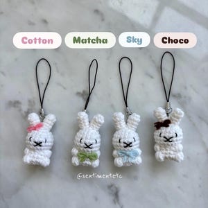 May include: Four white crochet bunny charms with black eyes and bows in pink, green, blue, and brown. Each bunny has a black cord for hanging. The text above the bunnies reads "Cotton", "Matcha", "Sky", and "Choco".