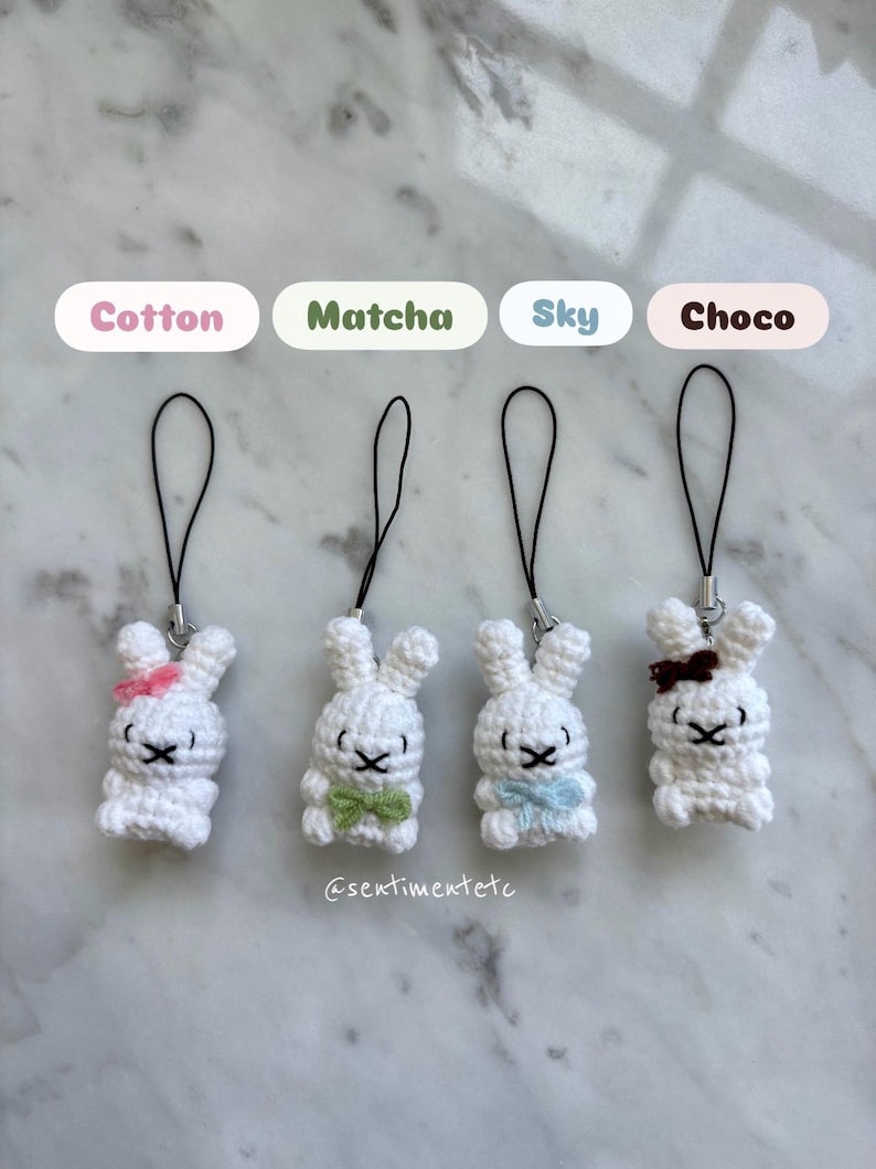 May include: Four white crochet bunny charms with black eyes and bows in pink, green, blue, and brown. Each bunny has a black cord for hanging. The text above the bunnies reads "Cotton", "Matcha", "Sky", and "Choco".