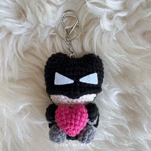 May include: A handmade crochet keychain featuring a black superhero figure with white eye accents and a pink heart. The keychain has a silver clasp and is set against a fluffy white background.