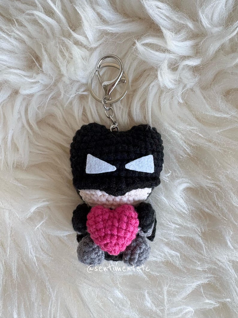 May include: A handmade crochet keychain featuring a black superhero figure with white eye accents and a pink heart. The keychain has a silver clasp and is set against a fluffy white background.