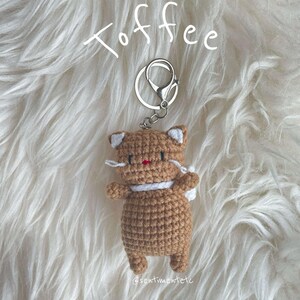 May include: A crocheted brown cat keychain with white accents, wearing a white scarf. The keychain is attached to a silver key ring and the word "Toffee" is written above the keychain. The keychain is on a white fluffy surface.