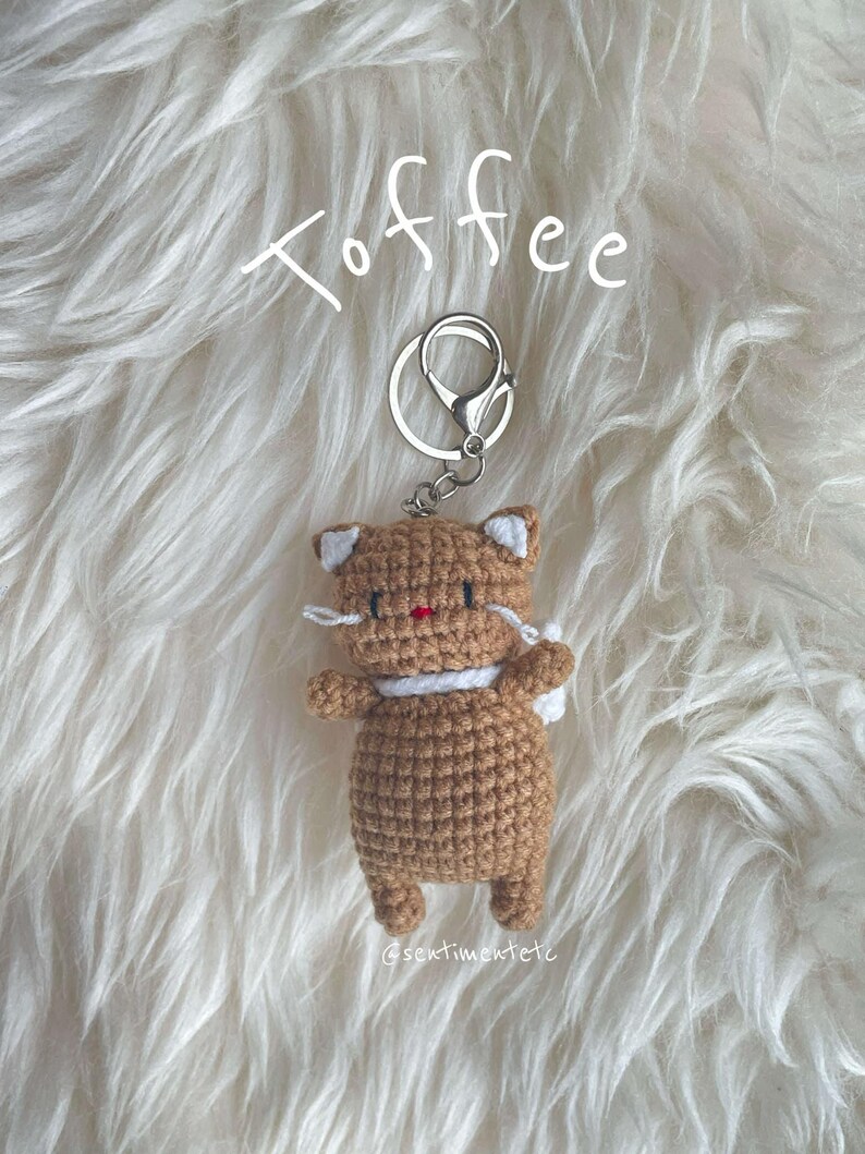 May include: A crocheted brown cat keychain with white accents, wearing a white scarf. The keychain is attached to a silver key ring and the word "Toffee" is written above the keychain. The keychain is on a white fluffy surface.