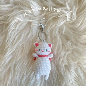 May include: White crocheted cat keychain with pink accents. The keychain is attached to a silver key ring. The text "Marshmallow" is written above the keychain.