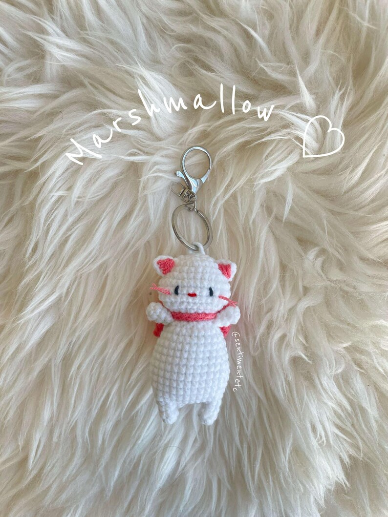 May include: White crocheted cat keychain with pink accents. The keychain is attached to a silver key ring. The text "Marshmallow" is written above the keychain.