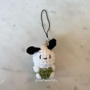 May include: A handmade crochet charm featuring a white body, black ears, and a green heart-shaped detail. The charm has a small, smiling face and a black strap for attachment. The text "@sentimentetc" is visible.