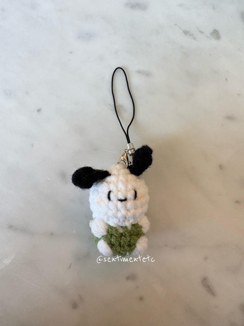 May include: A handmade crochet charm featuring a white body, black ears, and a green heart-shaped detail. The charm has a small, smiling face and a black strap for attachment. The text "@sentimentetc" is visible.