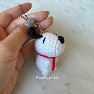 May include: A handmade crochet Snoopy charm. The white Snoopy has black ears and nose, with a red scarf. The charm is attached to a black string and a silver clasp. The Etsy shop name @sentimentetc is visible.