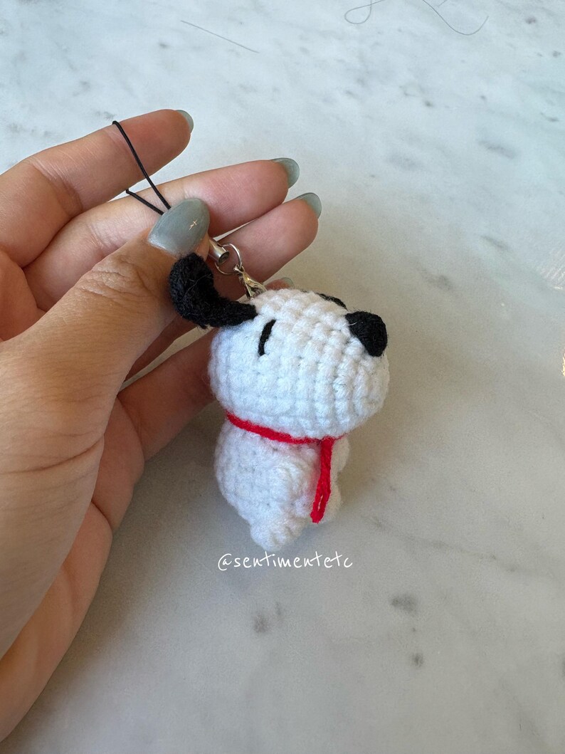 May include: A handmade crochet Snoopy charm. The white Snoopy has black ears and nose, with a red scarf. The charm is attached to a black string and a silver clasp. The Etsy shop name @sentimentetc is visible.