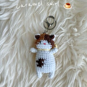 May include: A crocheted cat keychain with brown, white, and orange fur. The keychain is attached to a silver key ring. The cat is lying on a white fluffy surface. The text "Caramel swirl" is written above the cat. A small image of a caramel pudding is to the right of the text.