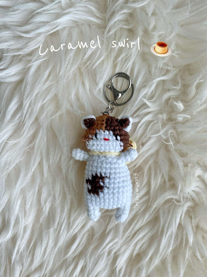 May include: A crocheted cat keychain with brown, white, and orange fur. The keychain is attached to a silver key ring. The cat is lying on a white fluffy surface. The text "Caramel swirl" is written above the cat. A small image of a caramel pudding is to the right of the text.