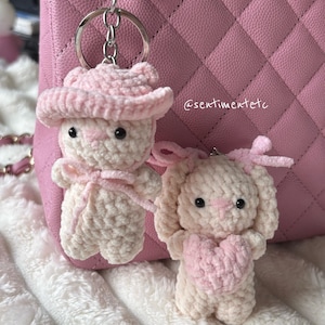 May include: Two handmade crochet bunny keychains. One bunny wears a pink cowboy hat and bow tie, the other holds a pink heart. Both are cream-colored with pink accents. The background includes a pink quilted purse and a fluffy white blanket.
